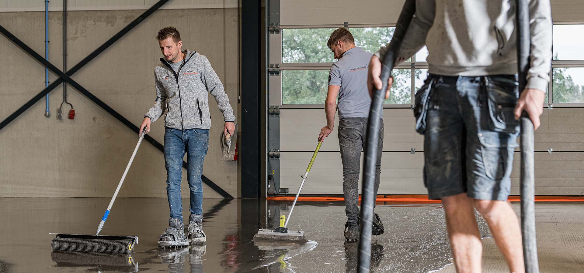 Werknemers brengen een beschermende toplaag aan op een betonnen vloer in een industriële ruimte, gebruikmakend van rollers en gereedschap