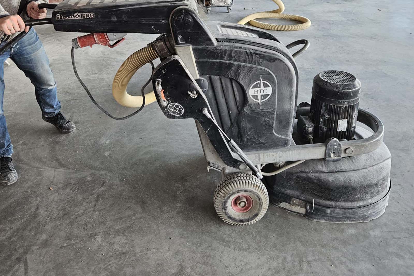 Een persoon bedient een vloerschuurmachine op een grijze betonvloer. De machine heeft een grote motor en stoffige onderdelen, met een slang voor het verzamelen van stof, perfect voor projecten zoals die op de Maasvlakte.