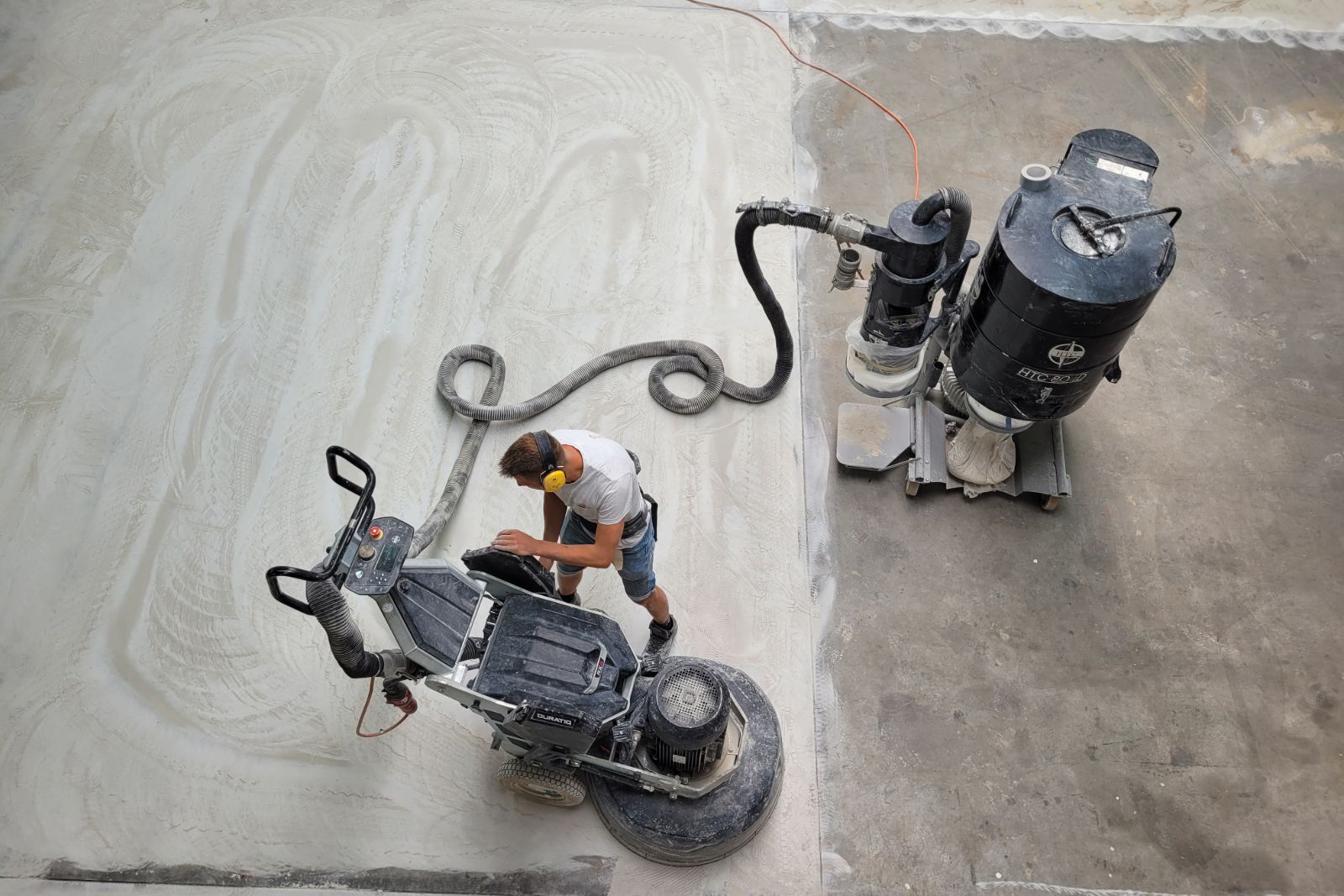 Man werkt met een machine aan een betonvloer die te hoog is gestort, waarbij het oppervlak wordt afgevlakt en stof wordt afgezogen.