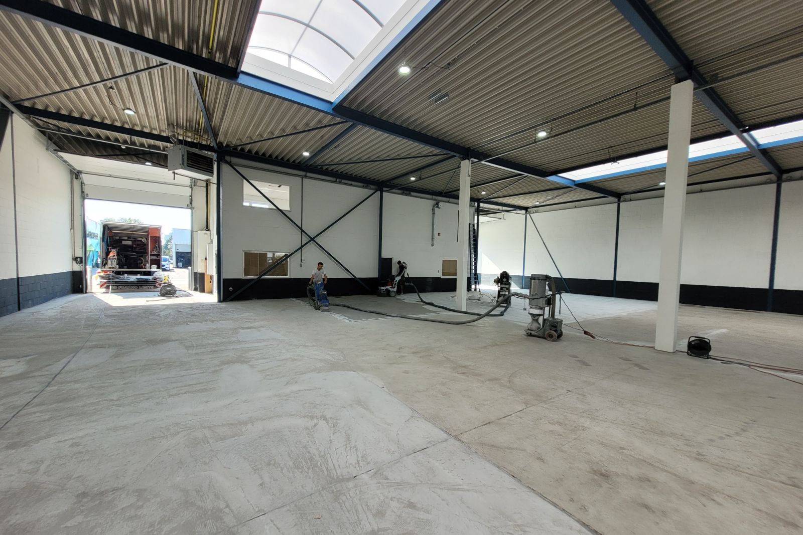 Overzicht van een loods waarin meerdere vakmensen een betonvloer die te hoog is gestort egaliseren met professionele vloerbewerkingsmachines.