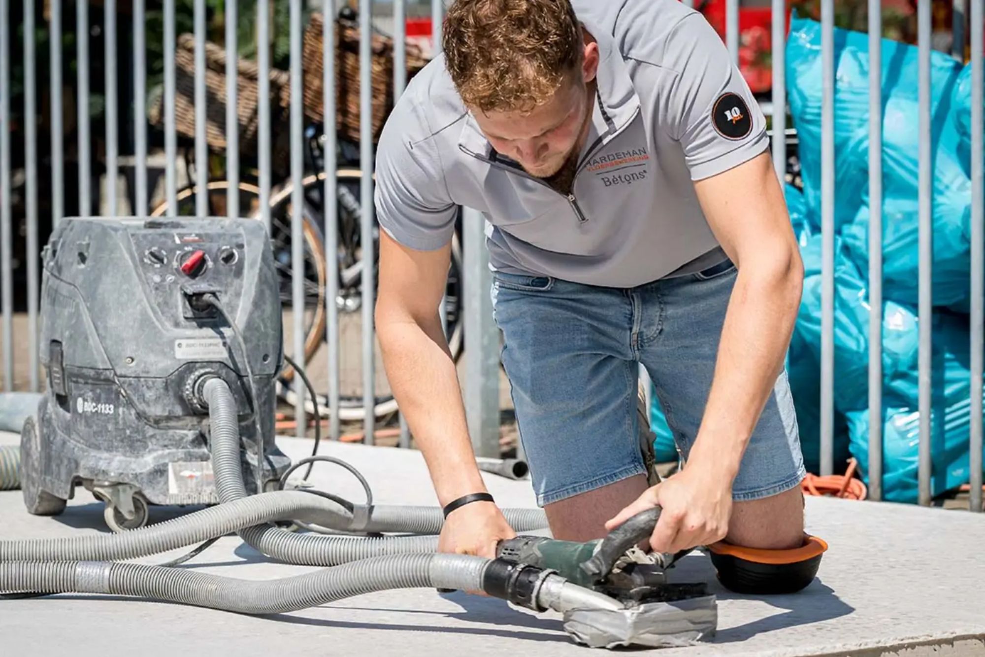 Vakman freest een betonvloer vlak met een industriële betonfreesmachine aangesloten op stofafzuiging. Voorgrond toont het gladde oppervlak, achtergrond met bouwmaterialen.